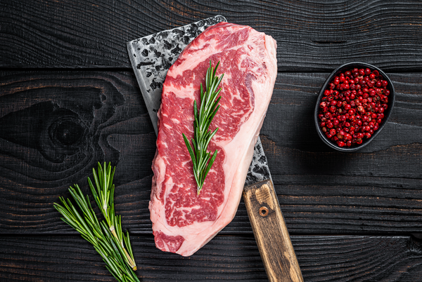 Raw New York Strip steak on dark wood background with a butchers knife and rosemary sprigs
